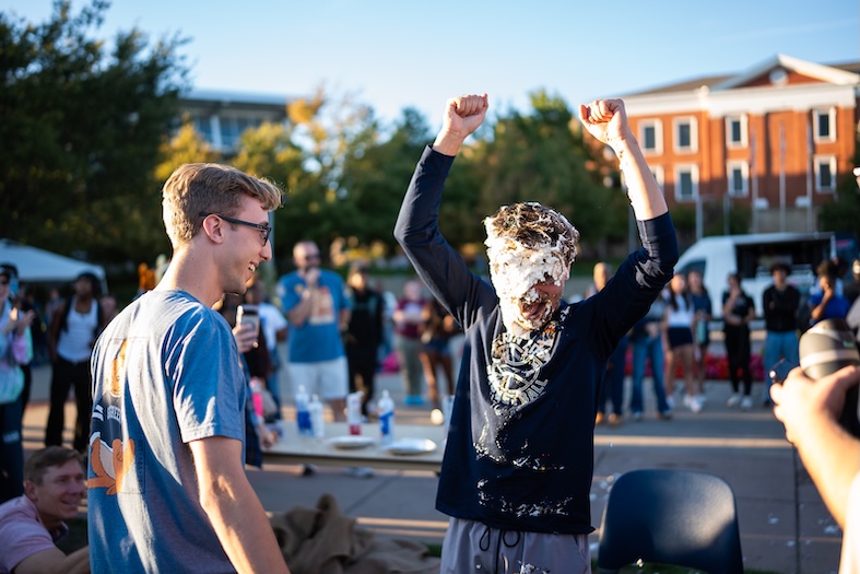 A person with their face covered in whipped cream raises their arms triumphantly while another person smiles nearby. A crowd watches the outdoor event on a sunny day, with tables and buildings in the background.